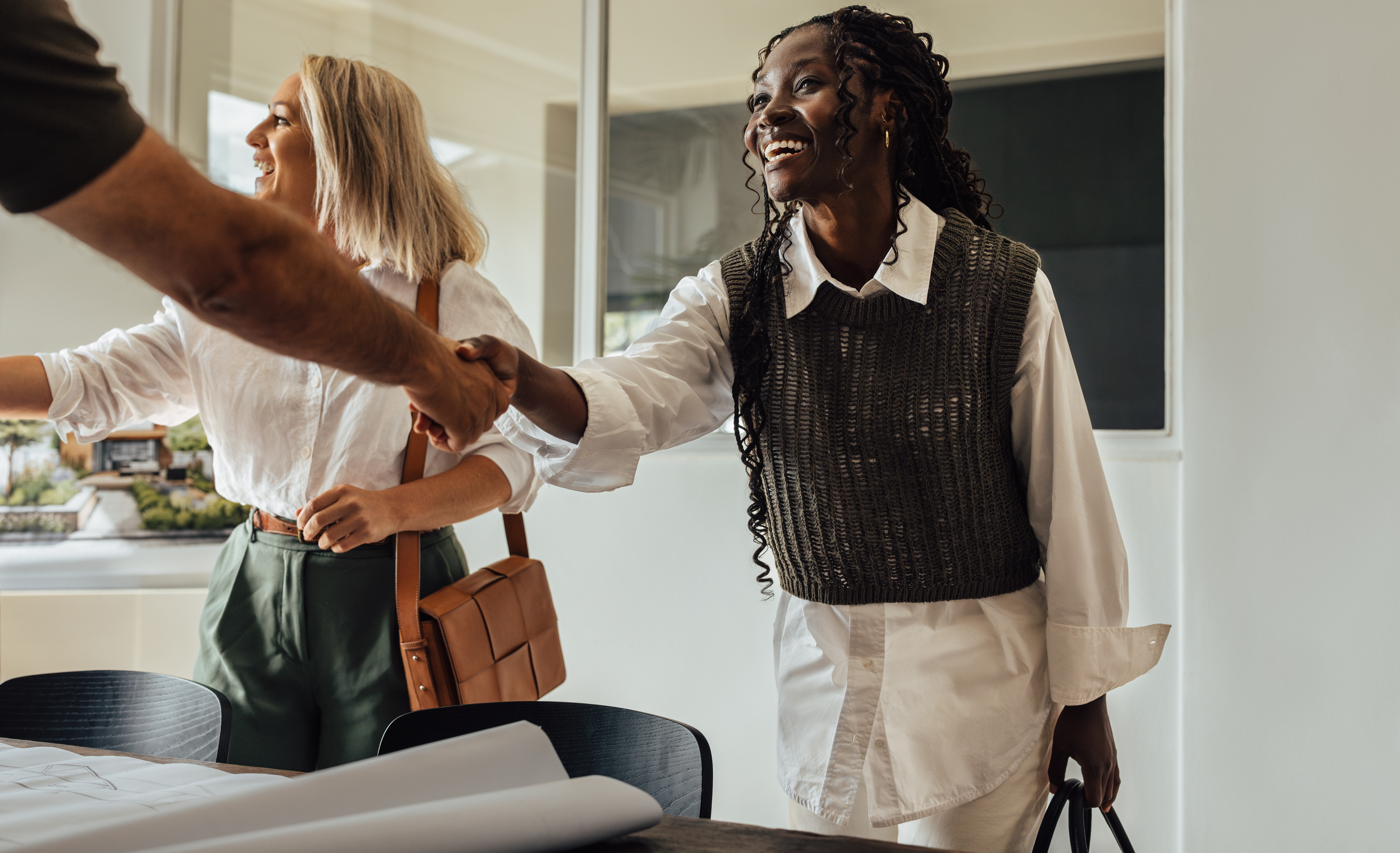 Two female architects, one young adult and one middle-aged, shake hands with a client in their modern office, symbolizing teamwork and professional collaboration.
